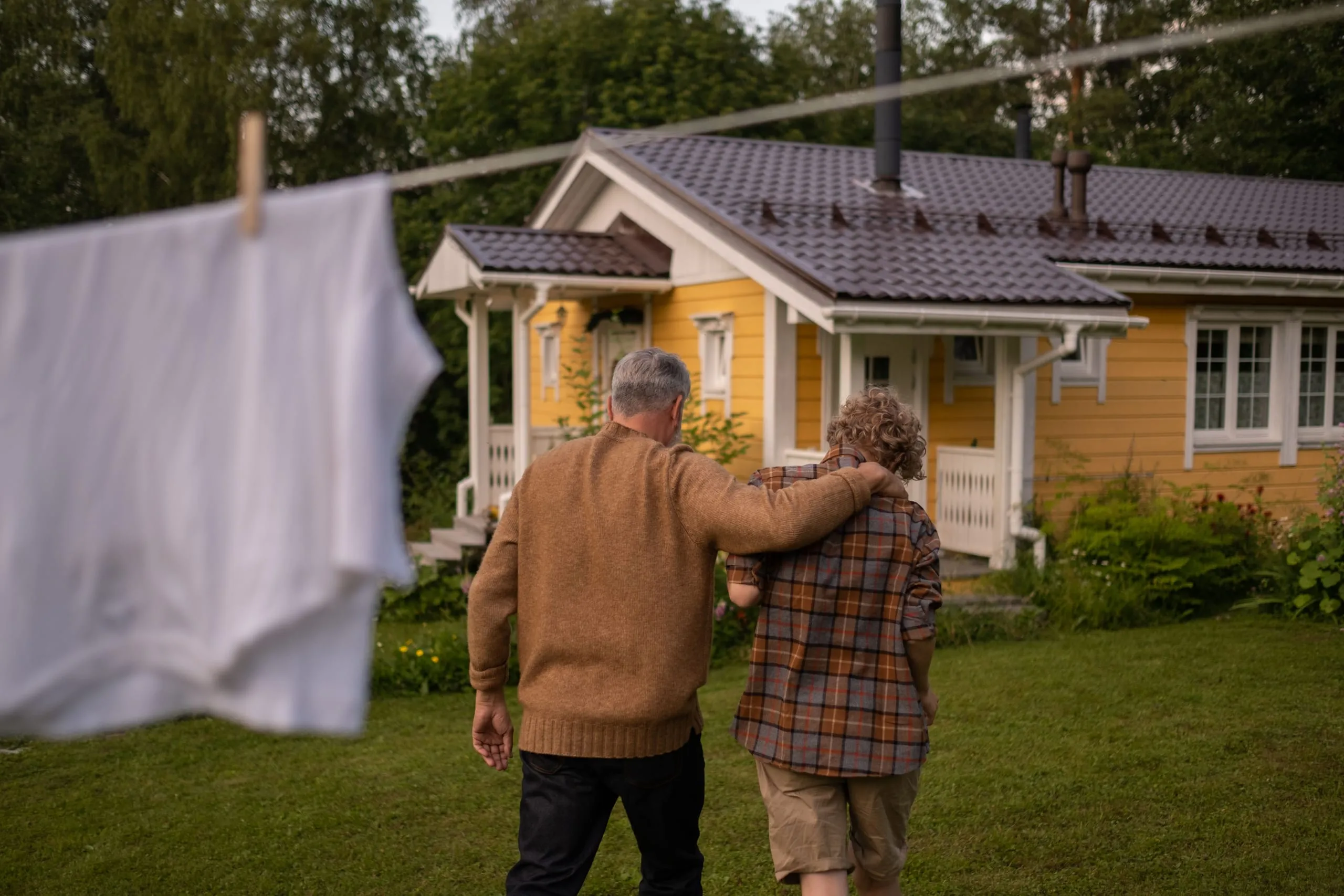 Isä ja lapsi kävelevät pyykkinarun alta kohti keltaista taloa.